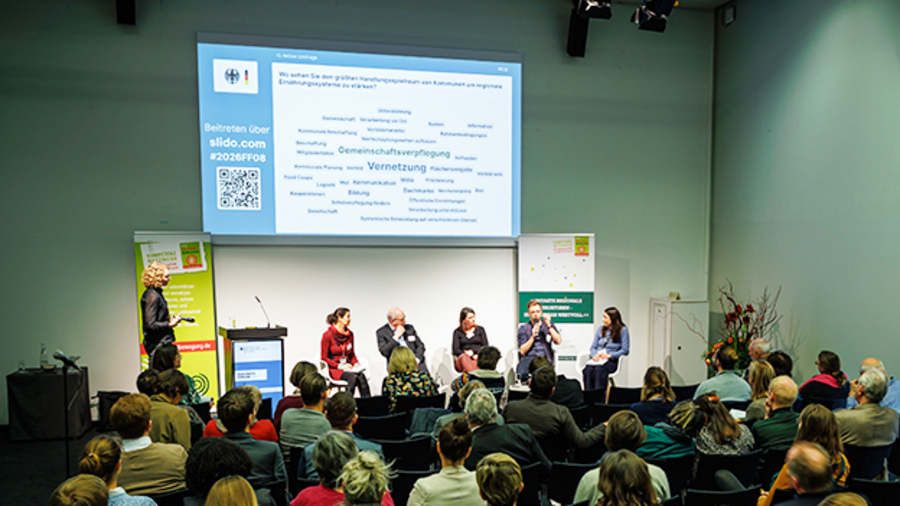 Das Podium zu einem der Fachforen mit Präsentation auf einer Leinwand im Hintergrund und einer Moderatorin. Im Vordergrund sind Zuhörende 